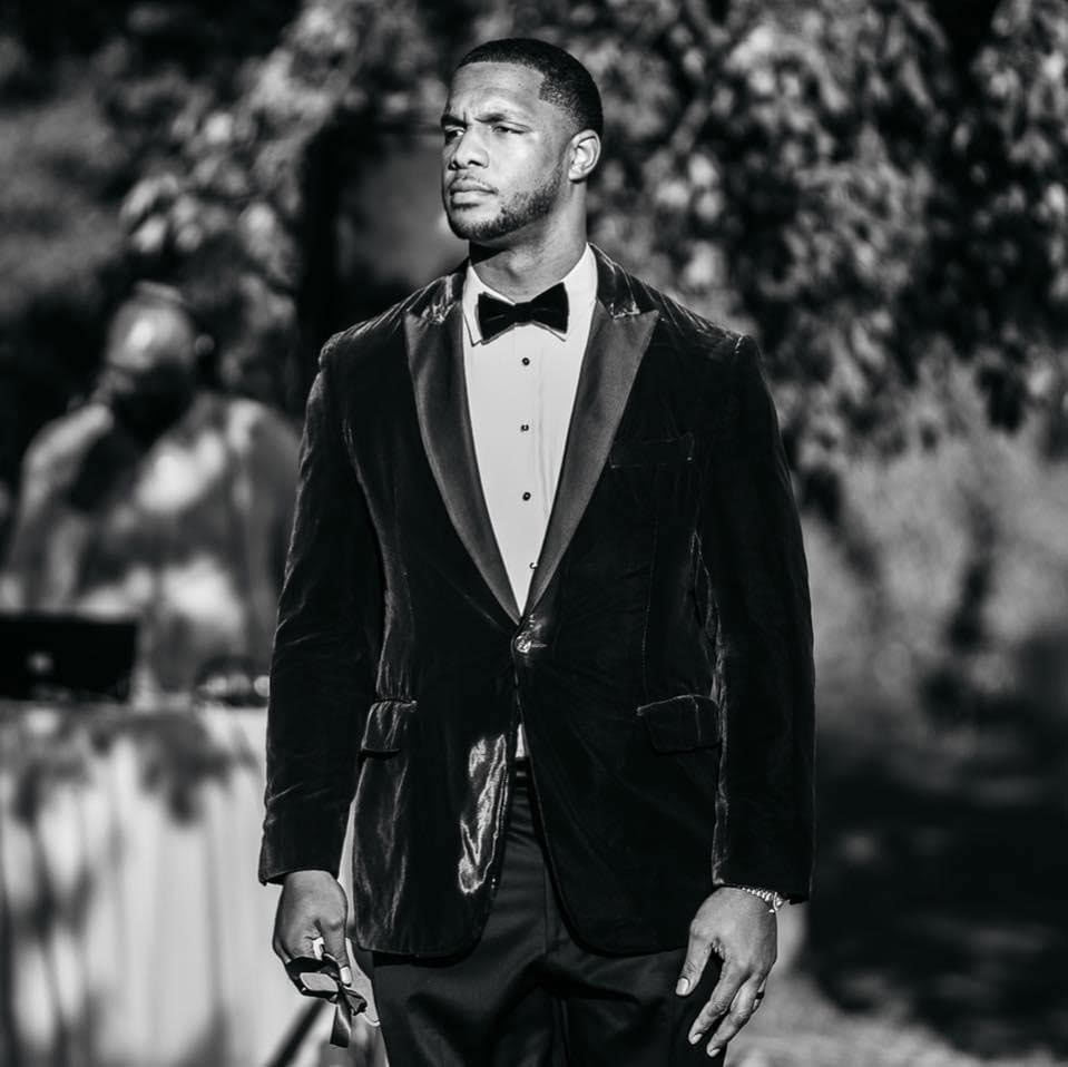 Quinton Coples in a tuxedo walking outdoors, holding sunglasses, in a black-and-white portrait.